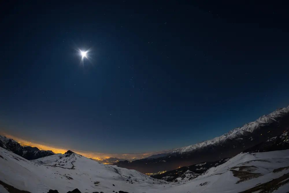 Night view of Turin city lights from snow-covered Alps by moonlight, with the Orion constellation and Sirius, the brightest star in the sky, visible in the fisheye shot.